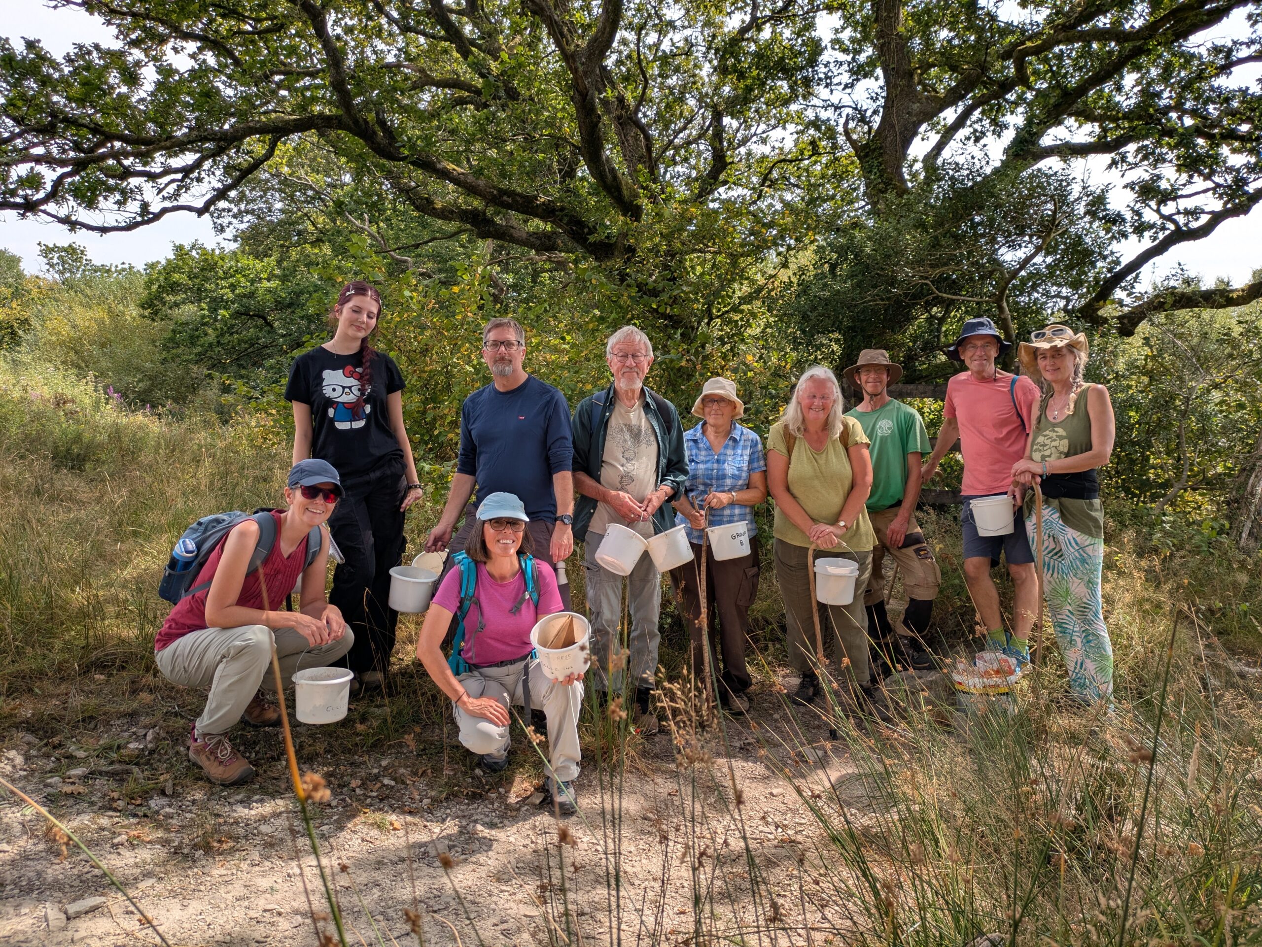Group of Moor Trees volunteers stood smiling in the shade of a tree on a sunny day with seed collecting buckets